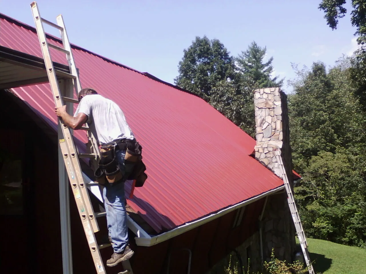 Roofer on a ladder beside a red metal roof and stone chimney — Soffit & Fascia Repair by Roofing Brothers in Monsey